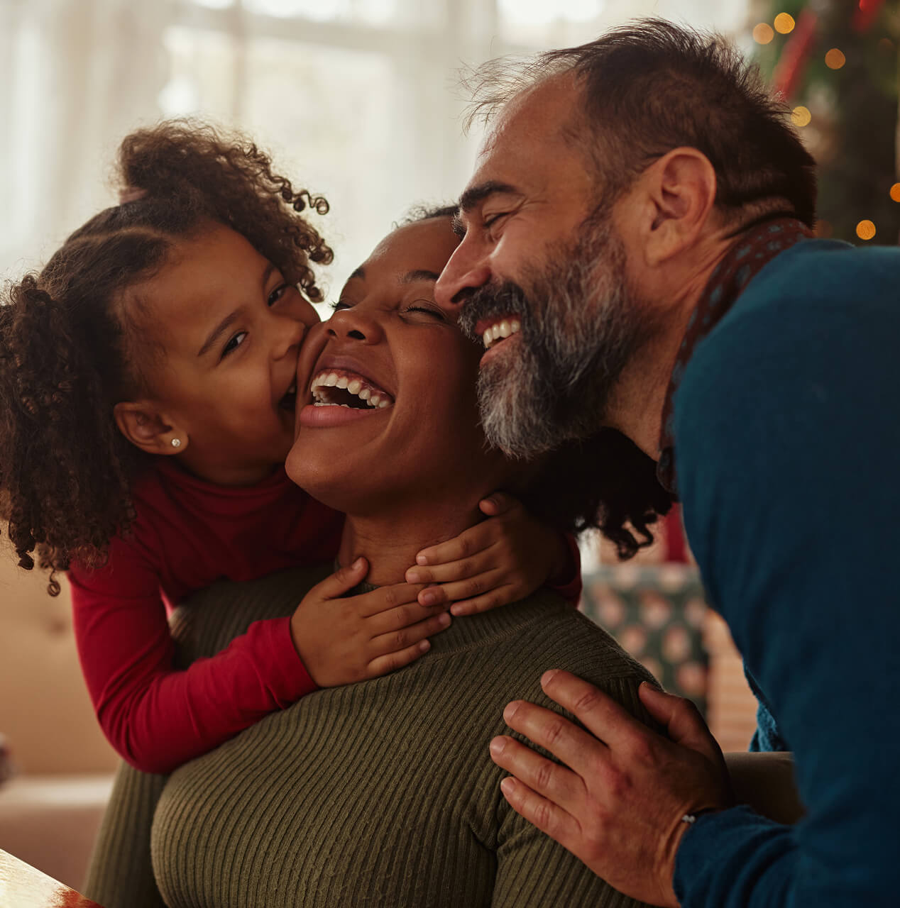 Family of three smiling and embracing in a warm indoor setting with a Christmas tree in the background.